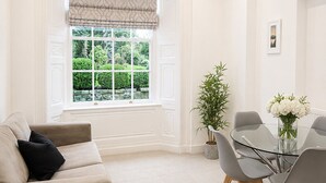 Ein modernes Wohnzimmer mit einem beigefarbenen Sofa, einem Glastisch mit Stühlen, einer Topfpflanze und einem Fenster mit Blick ins Grüne.
