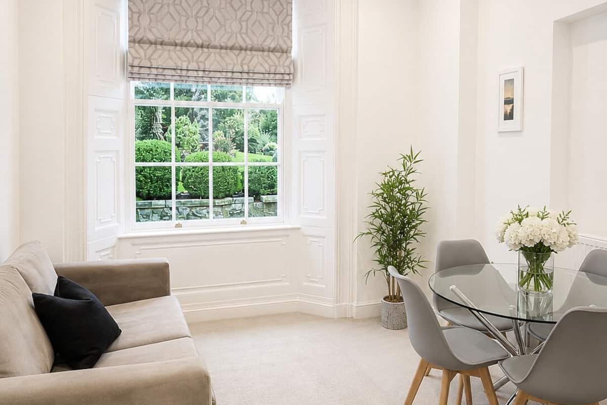 Un salon moderne avec un canapé beige, une table à manger en verre entourée de chaises, une plante en pot et une fenêtre donnant sur un paysage verdoyant.