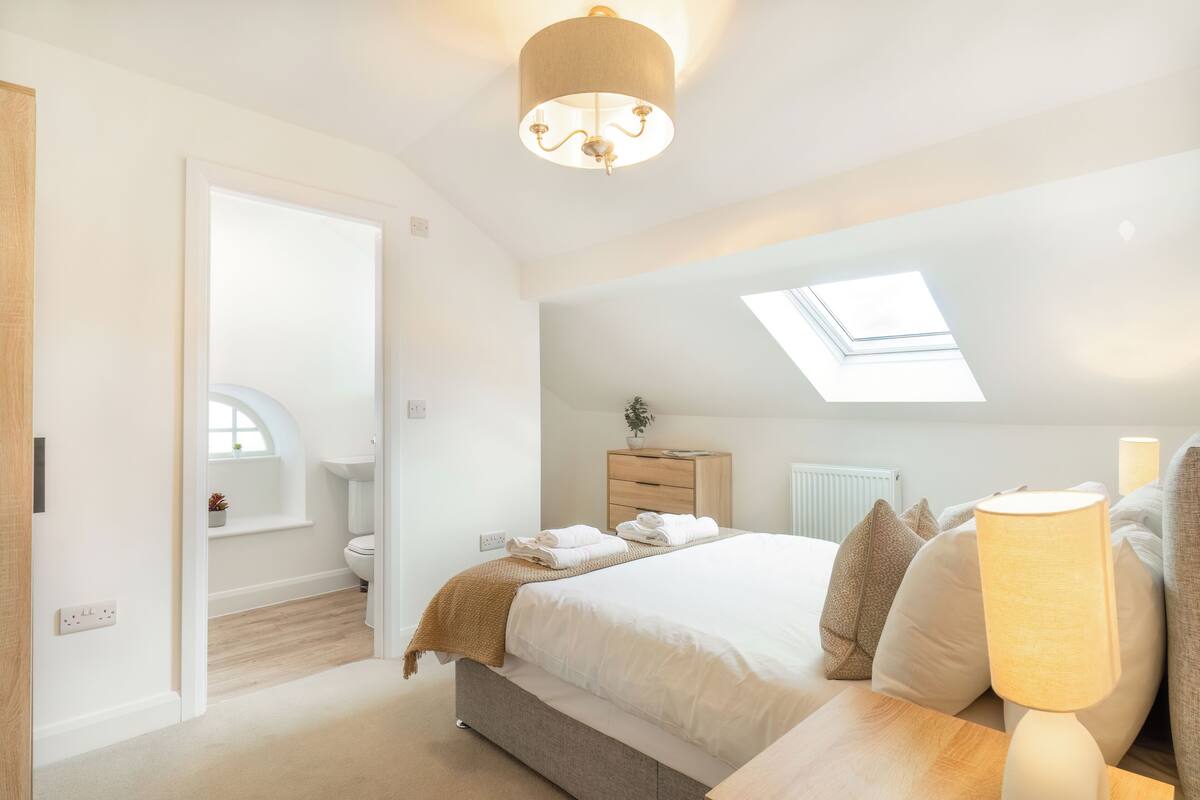 Une chambre avec un puits de lumière, une lampe de chevet, une table de chevet, un lit avec des oreillers, une commode, une fenêtre et une porte donnant sur une salle de bain.