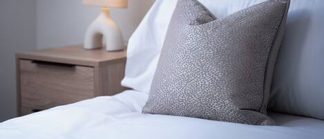 A neatly made bed with white linens and two grey pillows, a bedside table with a lamp, and a grey headboard.