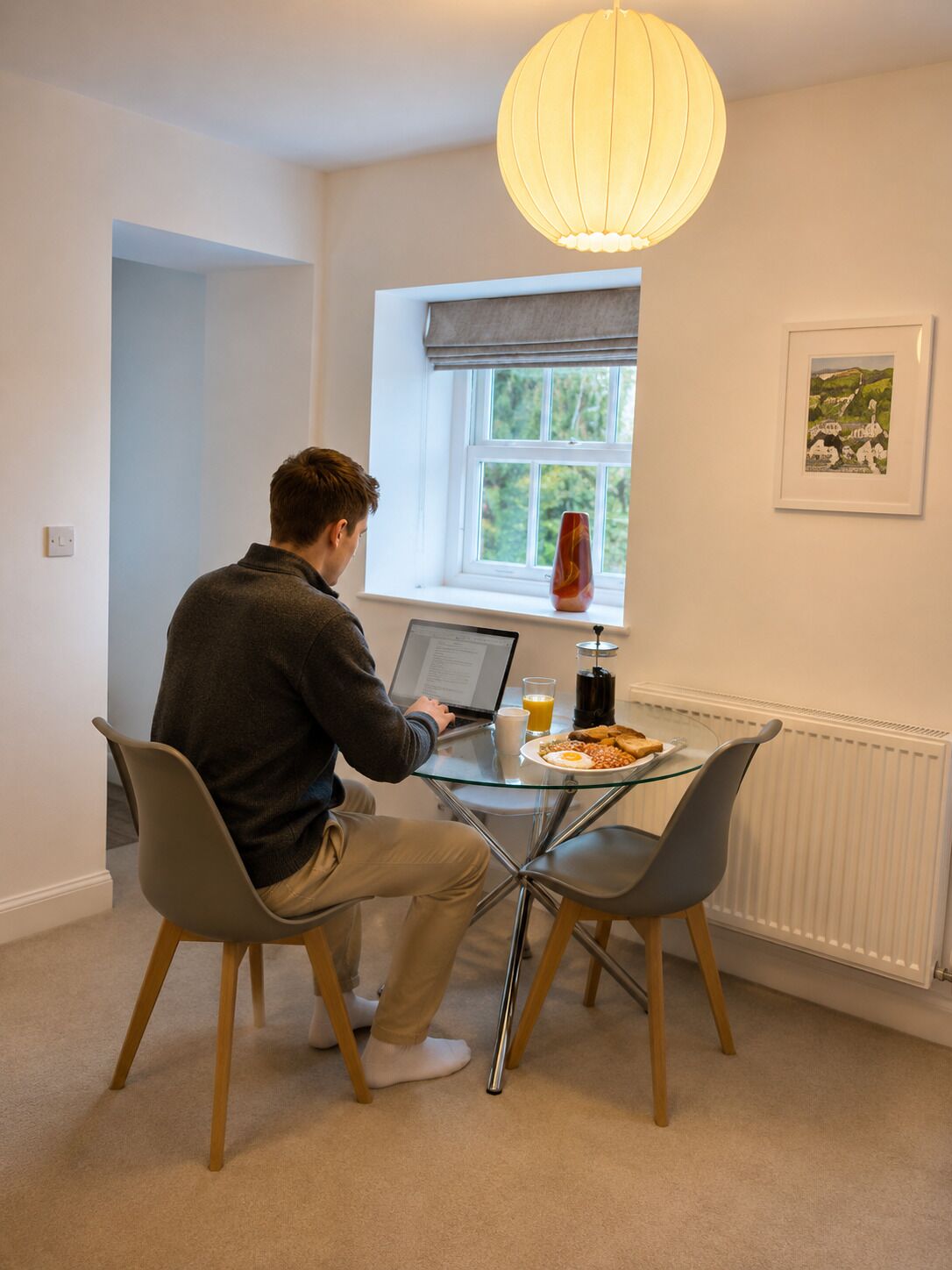 A person sitting at a glass table with a laptop, a plate of food, and a drink. There is a window with a view of greenery outside, a radiator, and a framed picture on the wall.