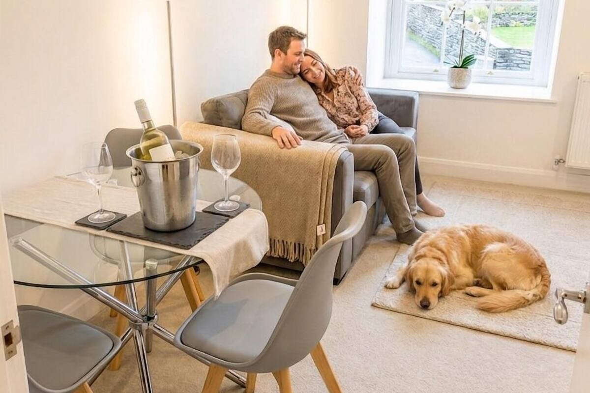 Un salon chaleureux avec un canapé, une table à manger et des chaises, un chien allongé sur le sol et une fenêtre donnant sur un paysage verdoyant.