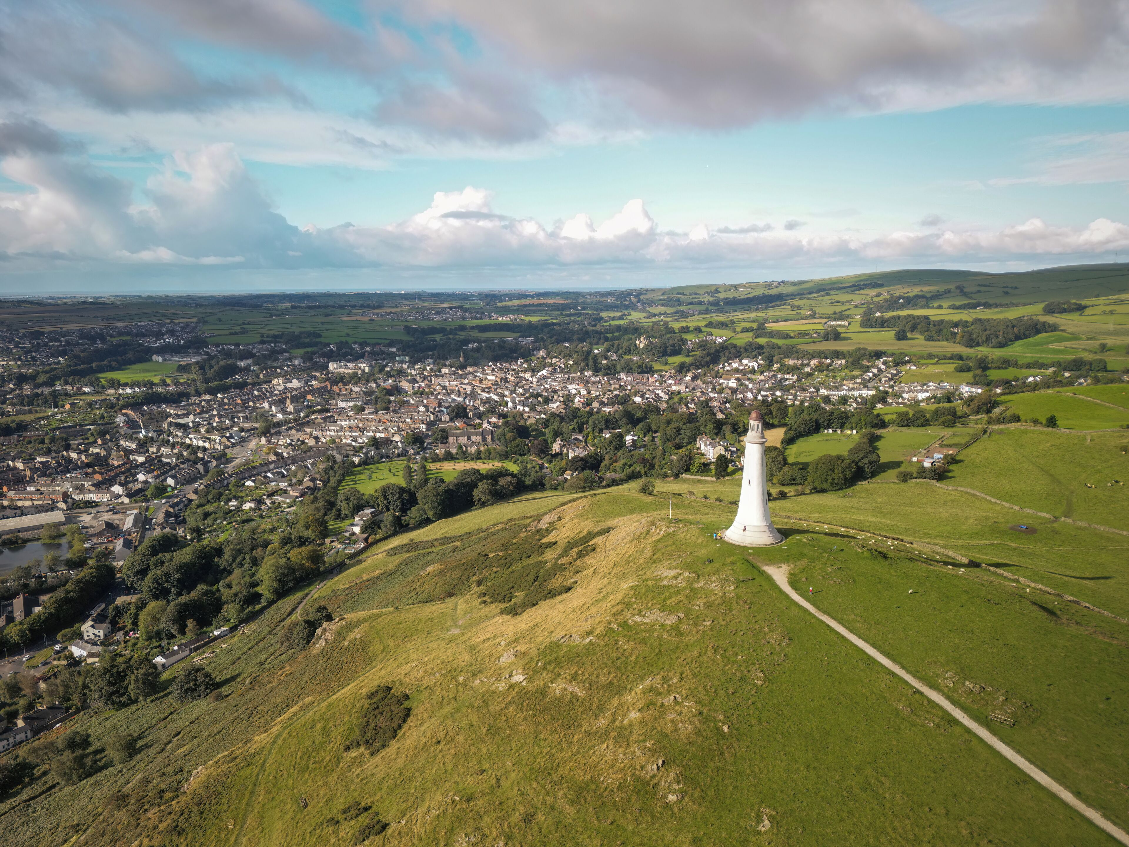 Un phare blanc, perché sur une colline, surplombe une ville entourée de champs verdoyants et sous un ciel dégagé.
