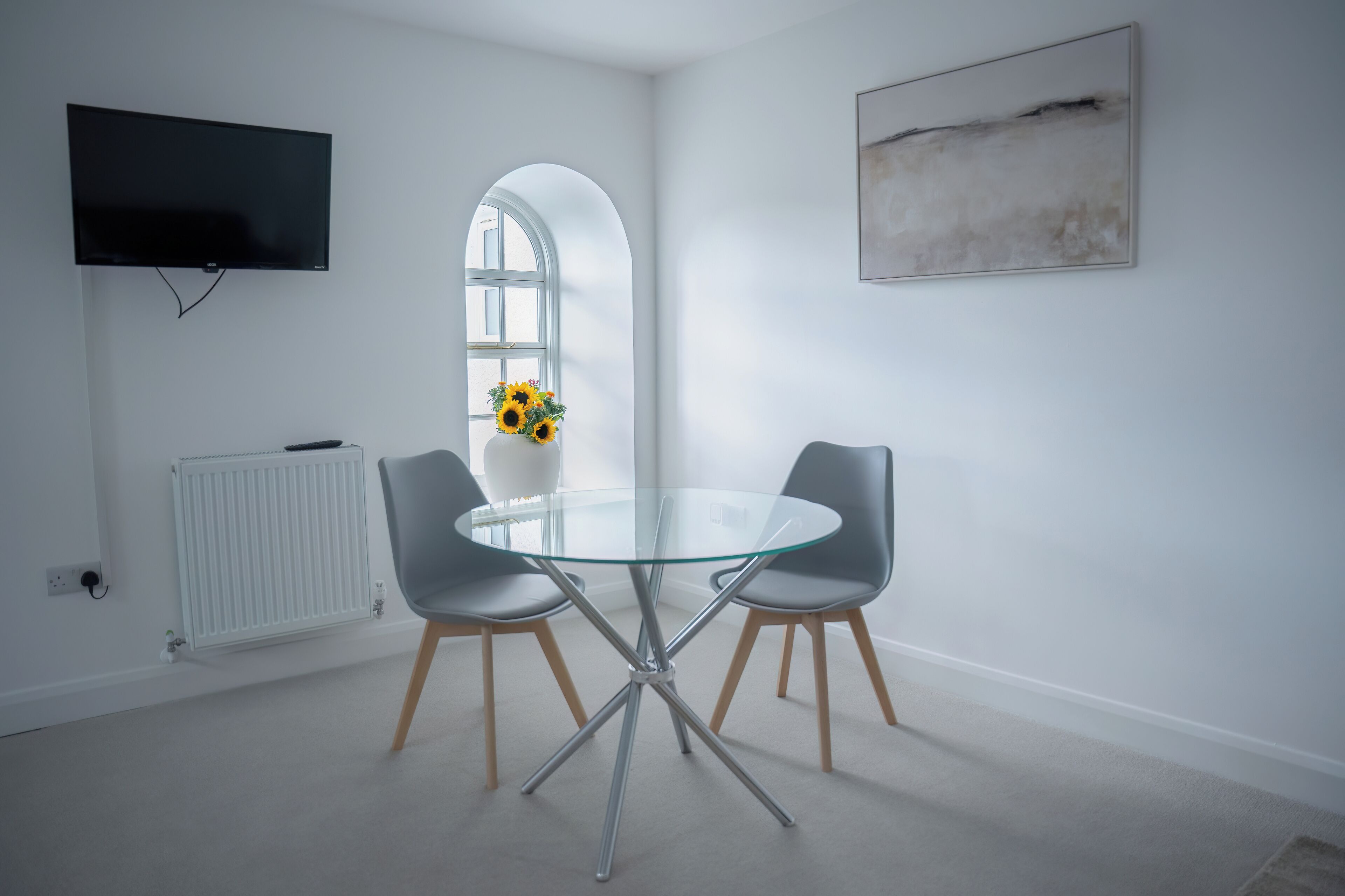 Une pièce moderne avec une table ronde en verre, deux chaises grises, un radiateur blanc, un téléviseur fixé au mur et une fenêtre agrémentée d’un vase avec des tournesols.