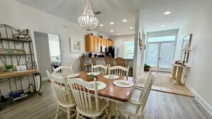 A dining area with a wooden table and white chairs, a chandelier, and a kitchen in the background. - Super Cute!   Easy Beach & Pool Access (Gulf Shores)