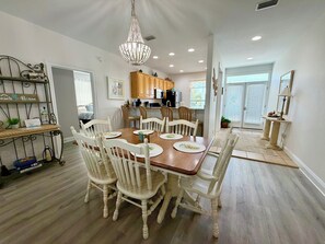 A dining area with a wooden table and white chairs, a chandelier, and a kitchen in the background.