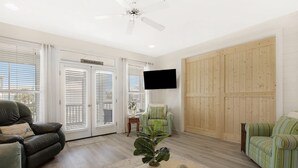 A spacious living room with a ceiling fan, a flat-screen TV mounted on the wall, and a large wooden door. - Super Cute!   Easy Beach & Pool Access (Gulf Shores)