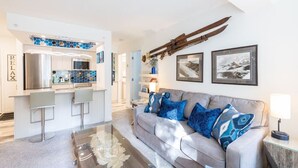 A modern living room with a grey sofa, blue patterned cushions, a glass coffee table, and a kitchen area with a blue hexagonal backsplash.