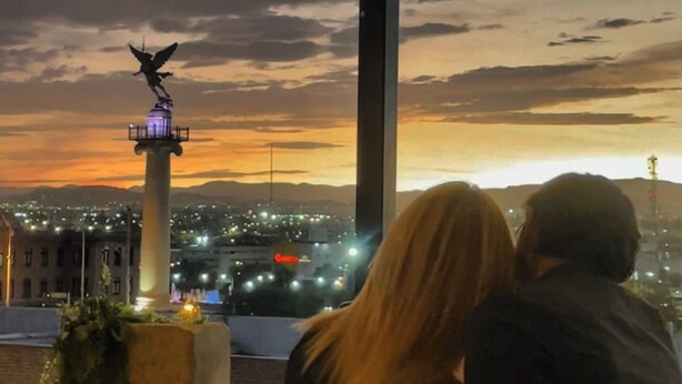 A couple seated at a table overlooking a cityscape with a prominent lighthouse and string lights. - AB Bed and breakfast  (Chihuahua)