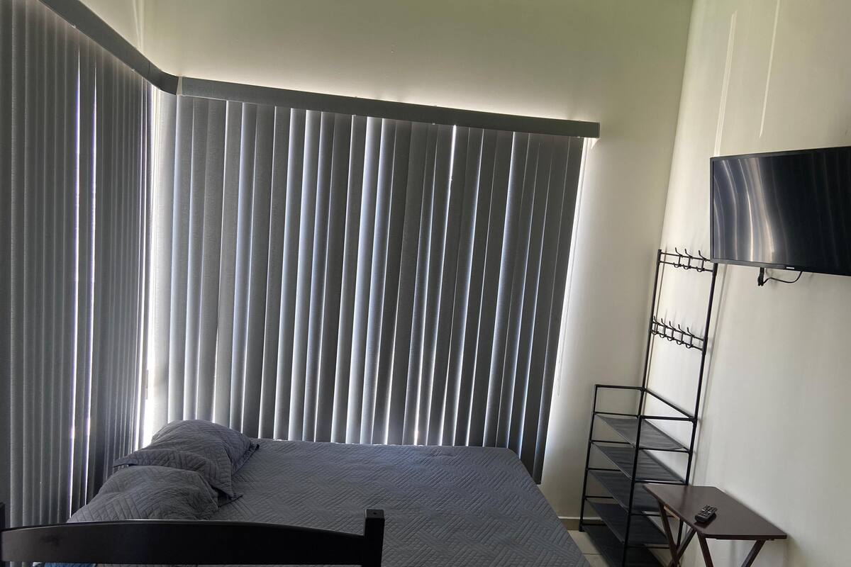 A bedroom with a bed, a TV mounted on the wall, and a window with vertical blinds.
