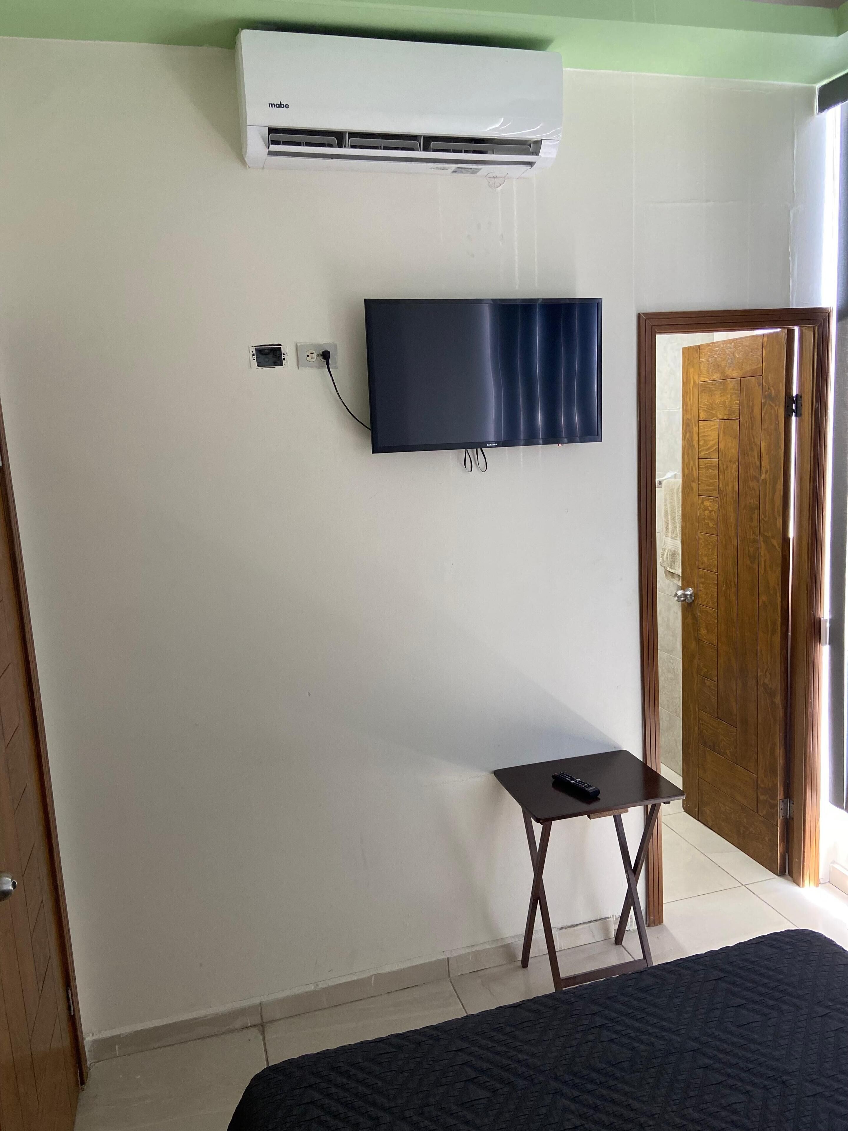 A bedroom with a bed, a small table, a television mounted on the wall, and an air conditioning unit above the TV.