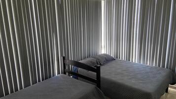 A room with two beds, each with a dark quilted bedspread, and a window with vertical blinds.