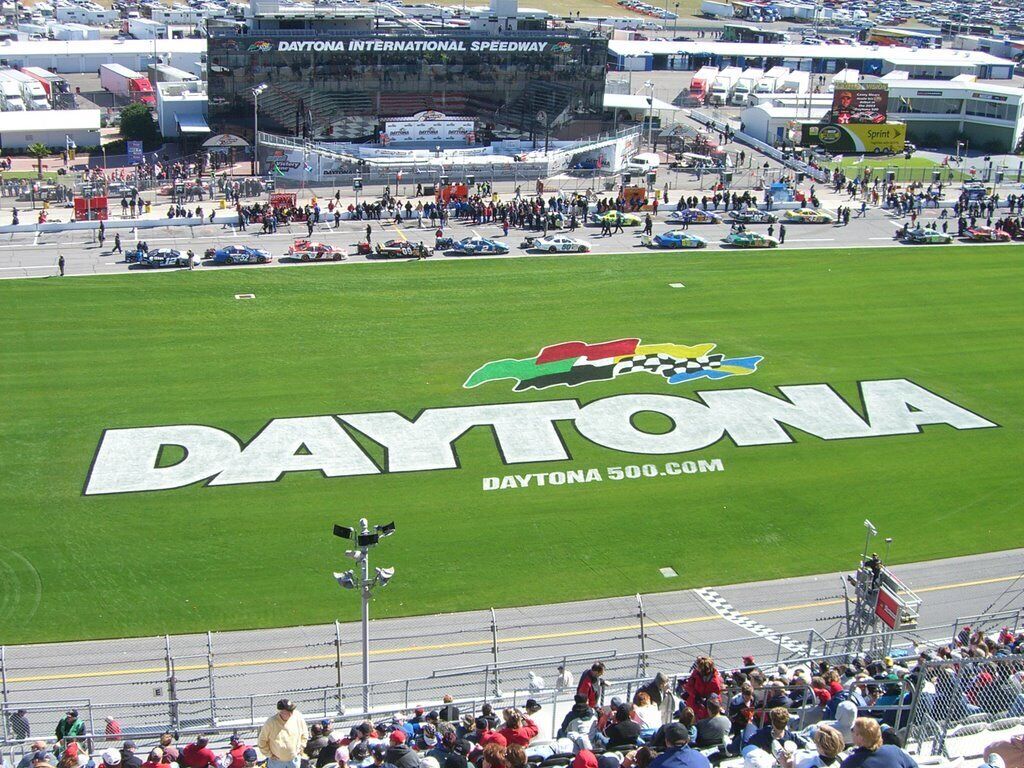 A stadium with a large "DAYTONA" logo on the field, surrounded by spectators and parked cars.