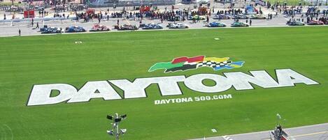 A stadium with a large "DAYTONA" logo on the field, surrounded by spectators and parked cars.