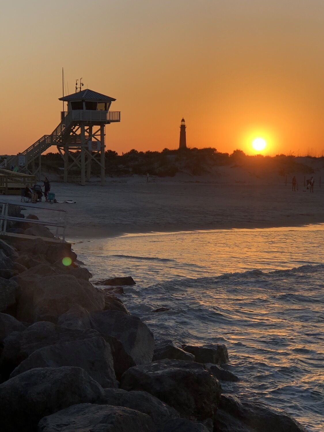 Eine Strandansicht mit einem Rettungsschwimmturm, einem Leuchtturm und der untergehenden Sonne über dem Meer.