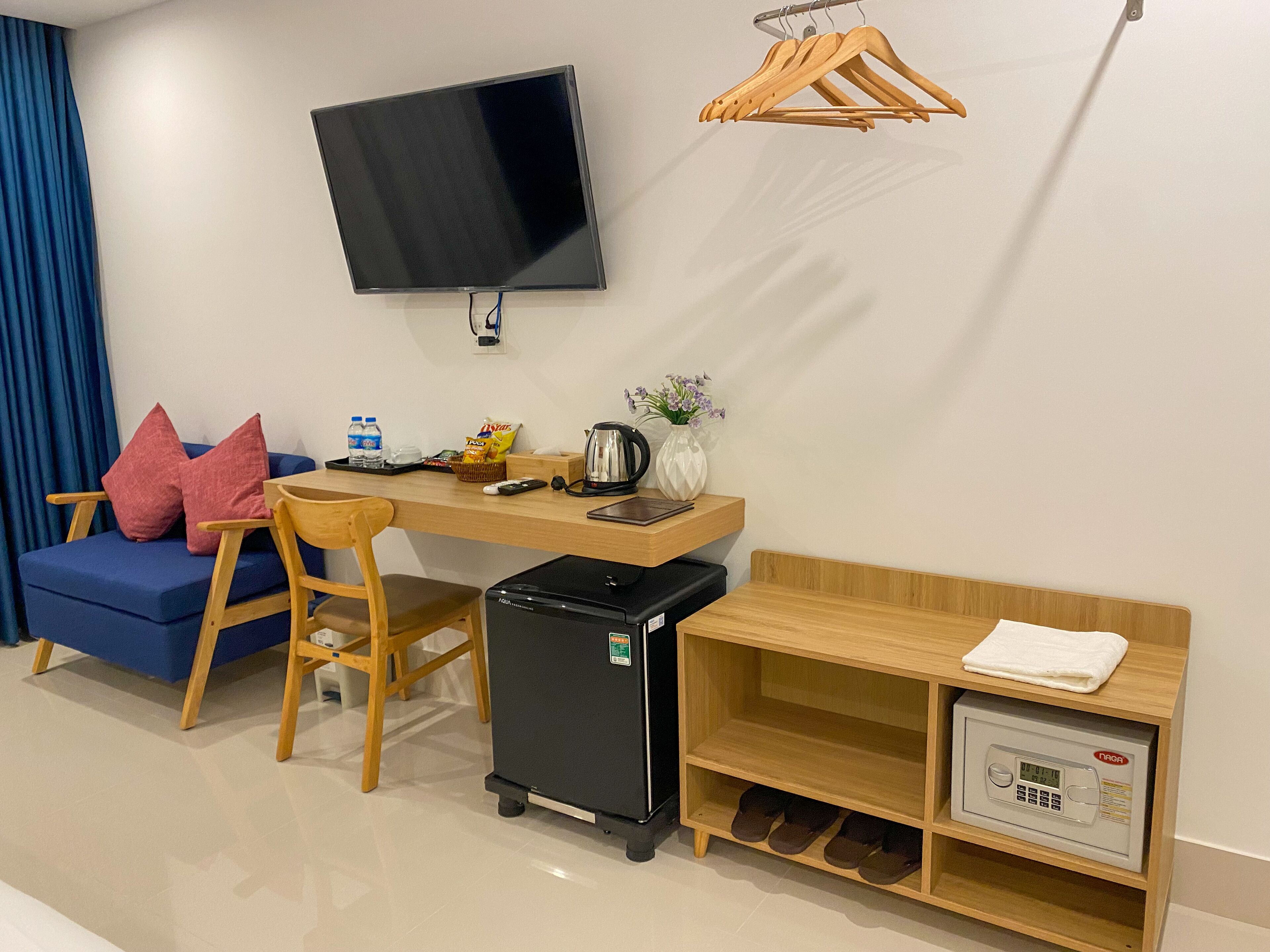 Une chambre d’hôtel compacte comprenant un bureau en bois, un petit réfrigérateur noir, un téléviseur fixé au mur, un canapé bleu avec des coussins roses et un porte-chaussures.