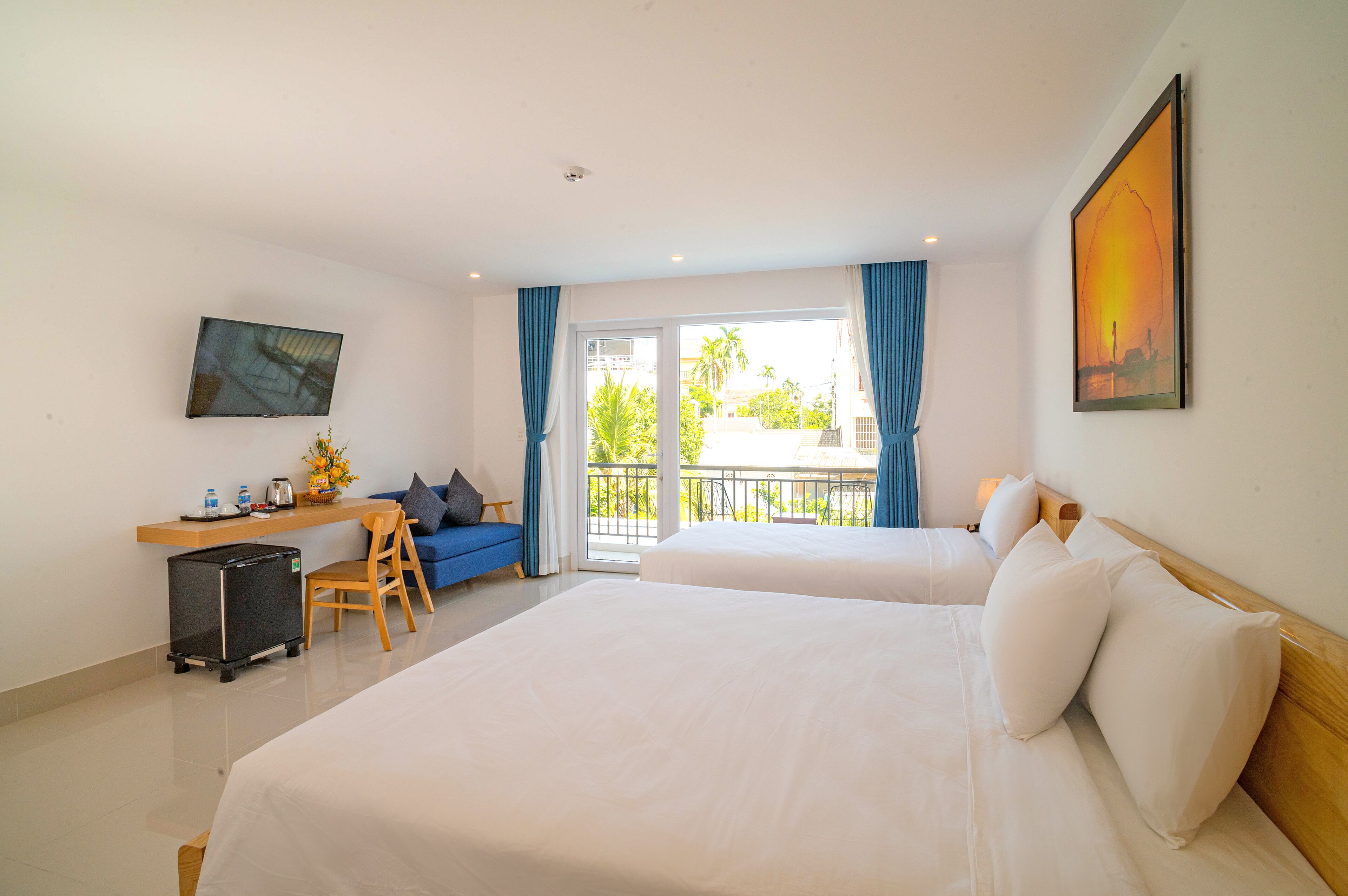 A hotel room with two beds, a desk, a chair, a small refrigerator, and a balcony with a view of palm trees.