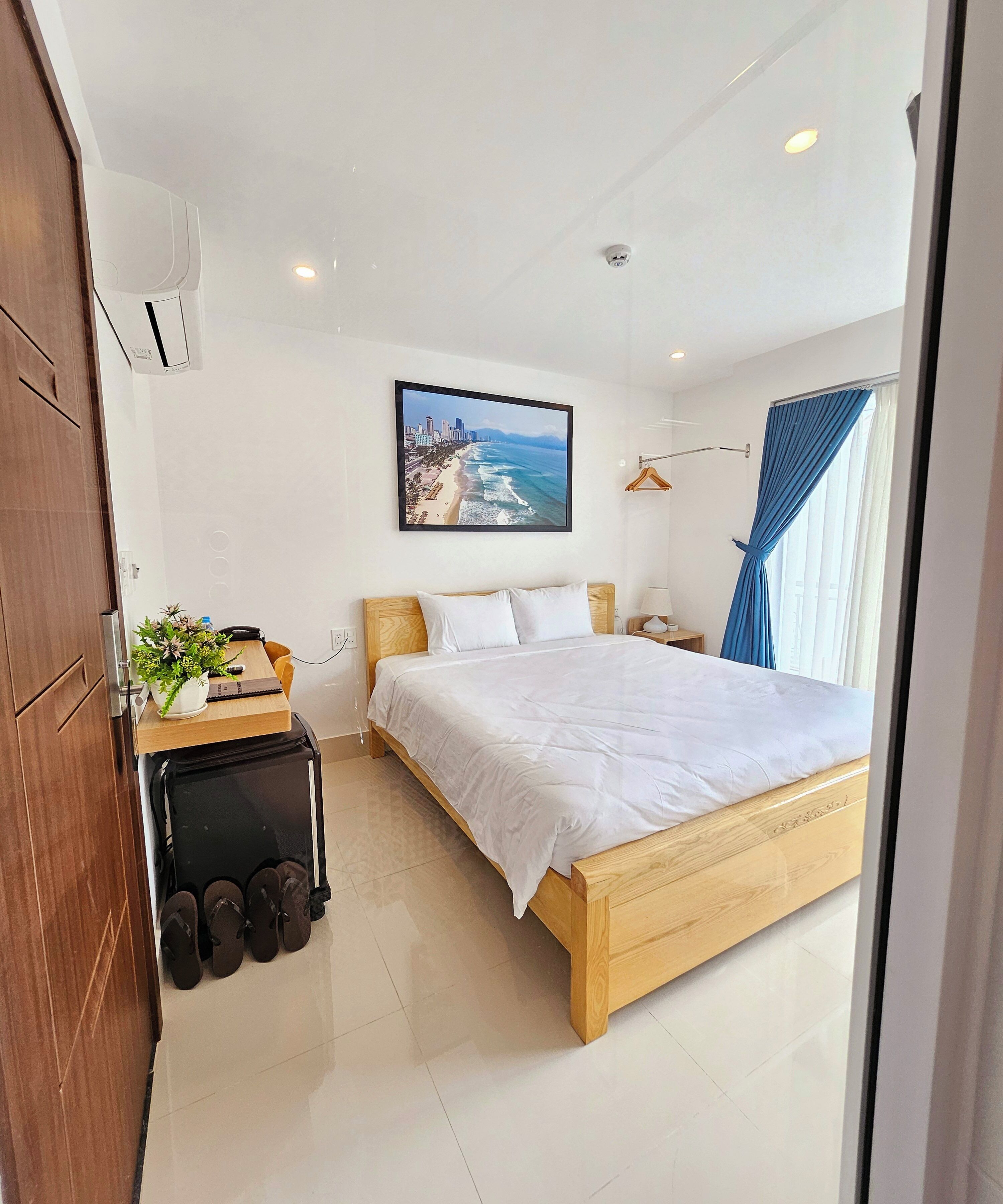 A modern hotel room with a wooden bed, a bedside table with a lamp, a wall-mounted air conditioner, and a framed picture of a beach.