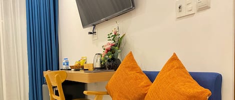Una habitación con un sofá azul, dos cojines naranjas, una silla de madera, un escritorio con una tetera, un florero y un televisor montado en la pared.
