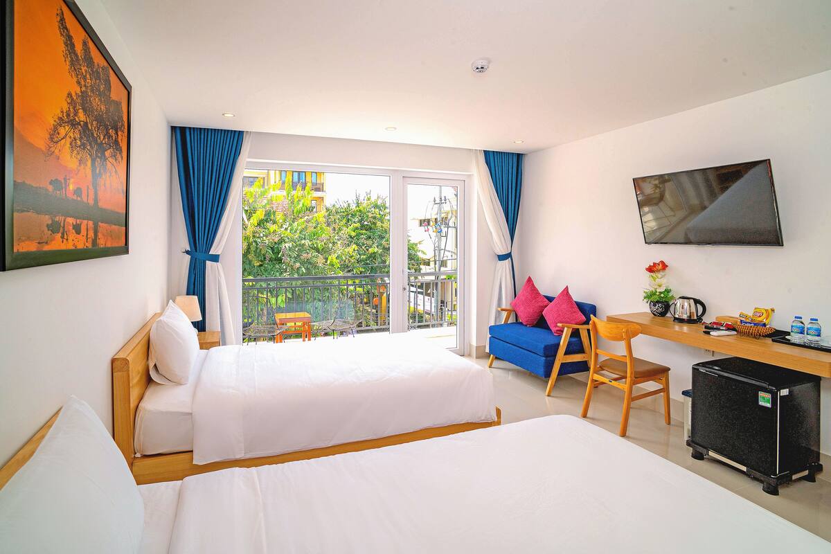 Ein Hotelzimmer mit zwei Betten, einem Schreibtisch mit Stuhl, einem kleinen Kühlschrank und einem Balkon mit Blick ins Grüne.