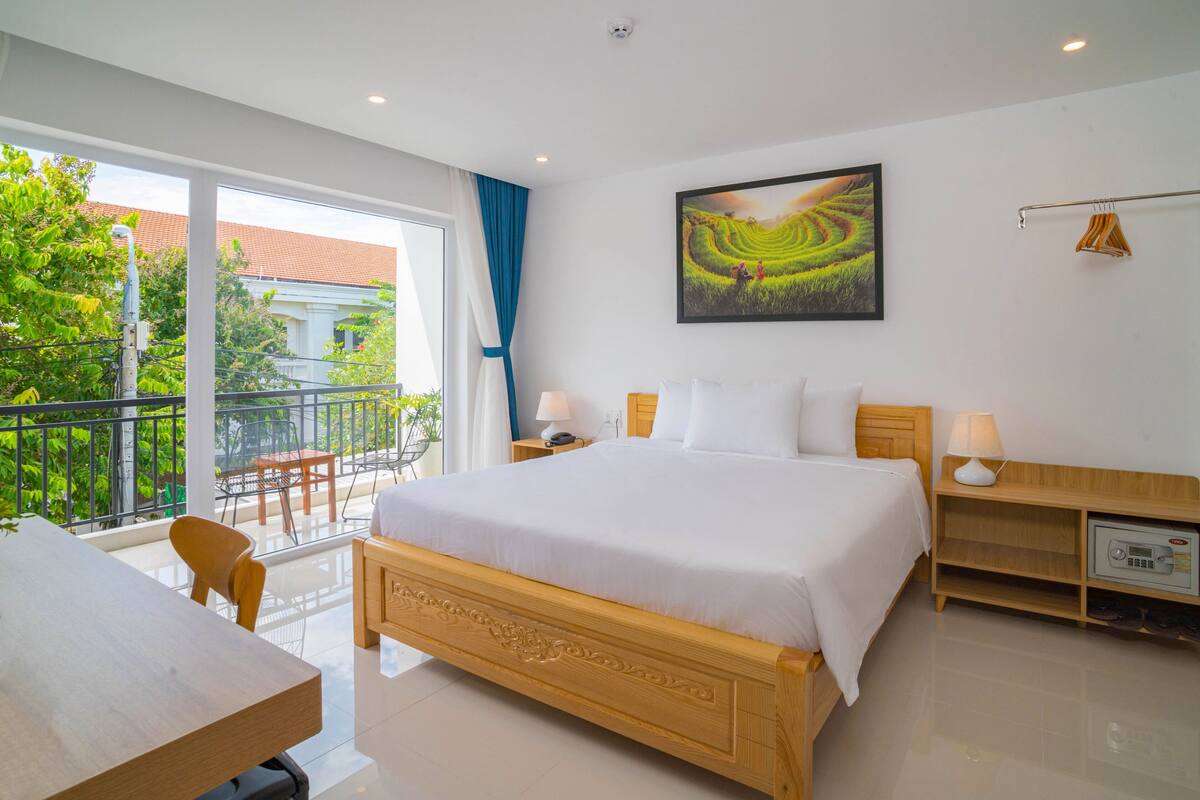 Ein Hotelzimmer mit einem großen Bett, einem Schreibtisch und einem Balkon mit Blick auf Grünflächen und ein Gebäude.