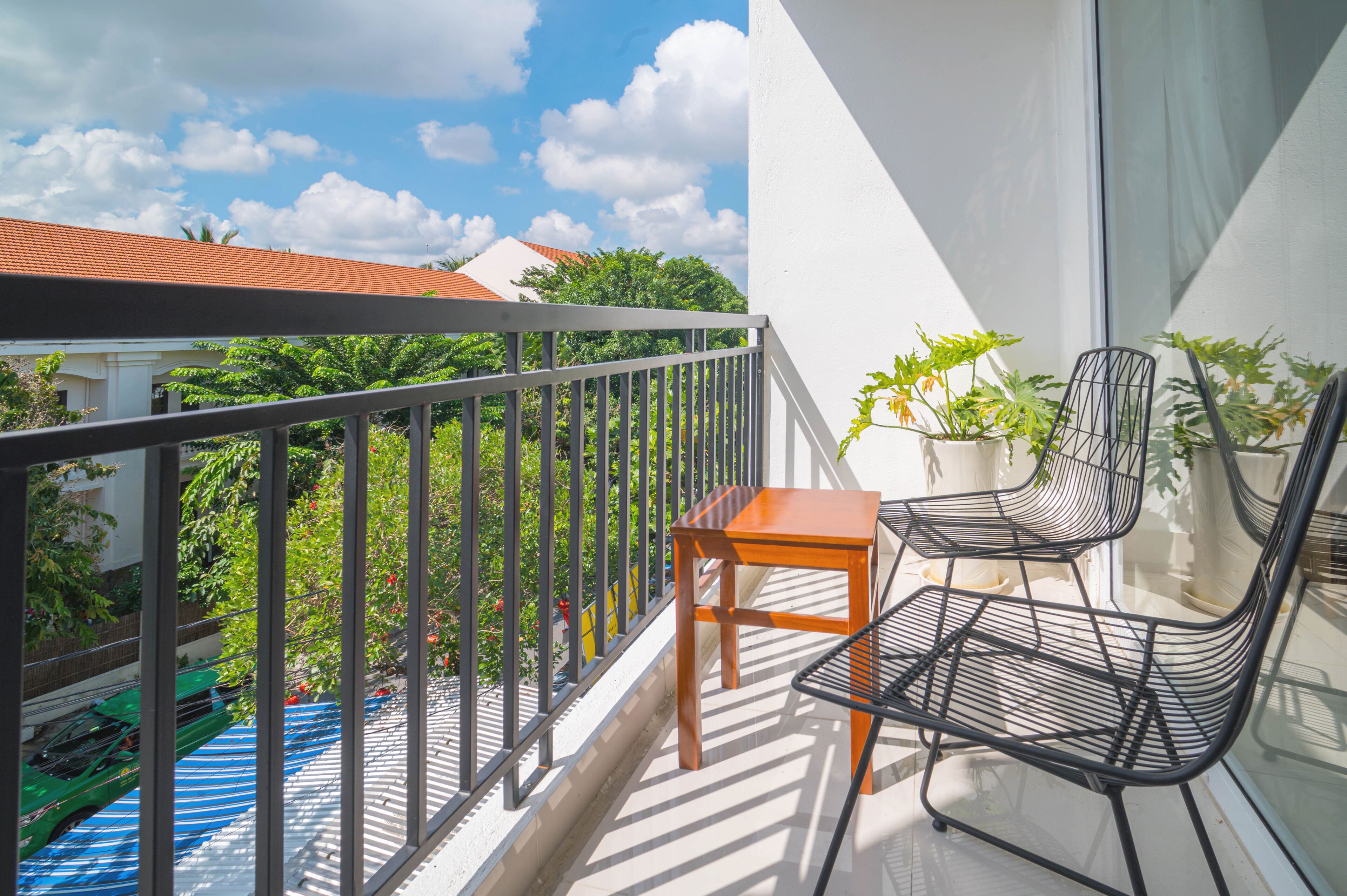 Un balcon doté d’une rambarde en métal, d’une table en bois et de deux chaises en fil de fer noir. Des plantes en pot sont disposées sur le balcon.