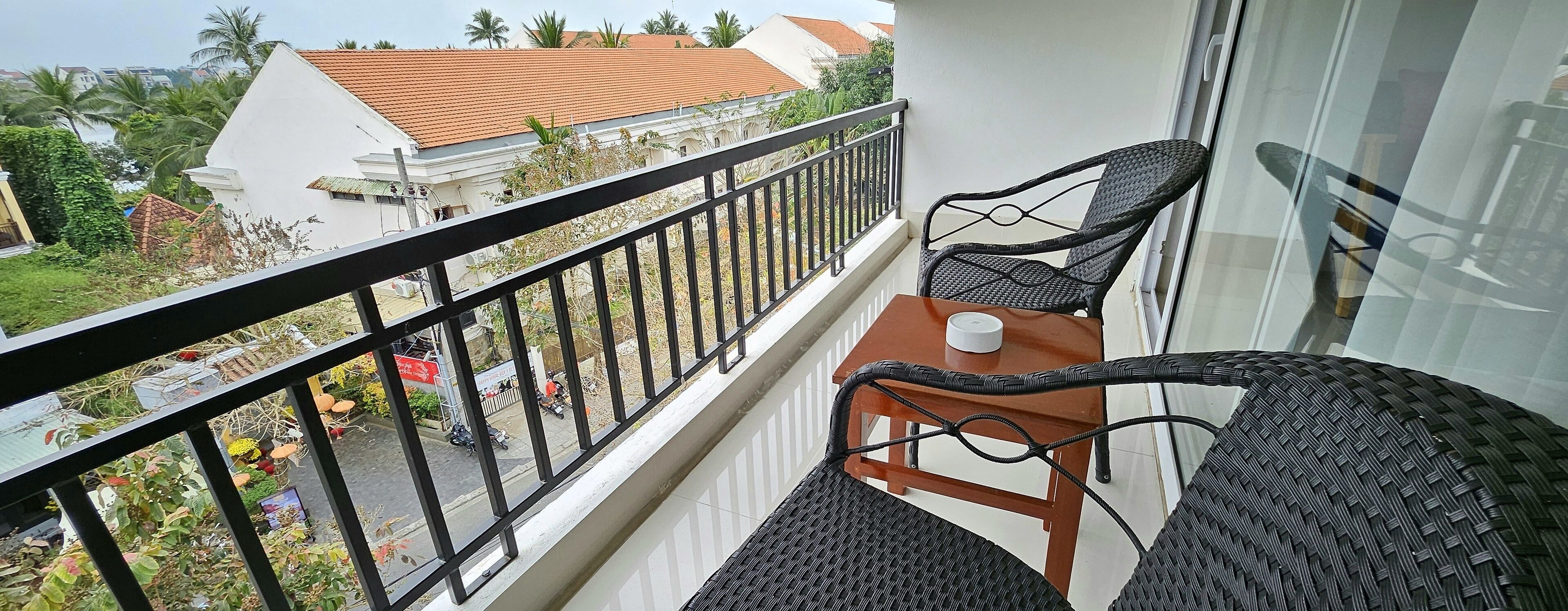 Ein Balkon mit Korbmöbeln und einem kleinen Tisch, mit Blick auf eine Straße mit Gebäuden und Palmen.