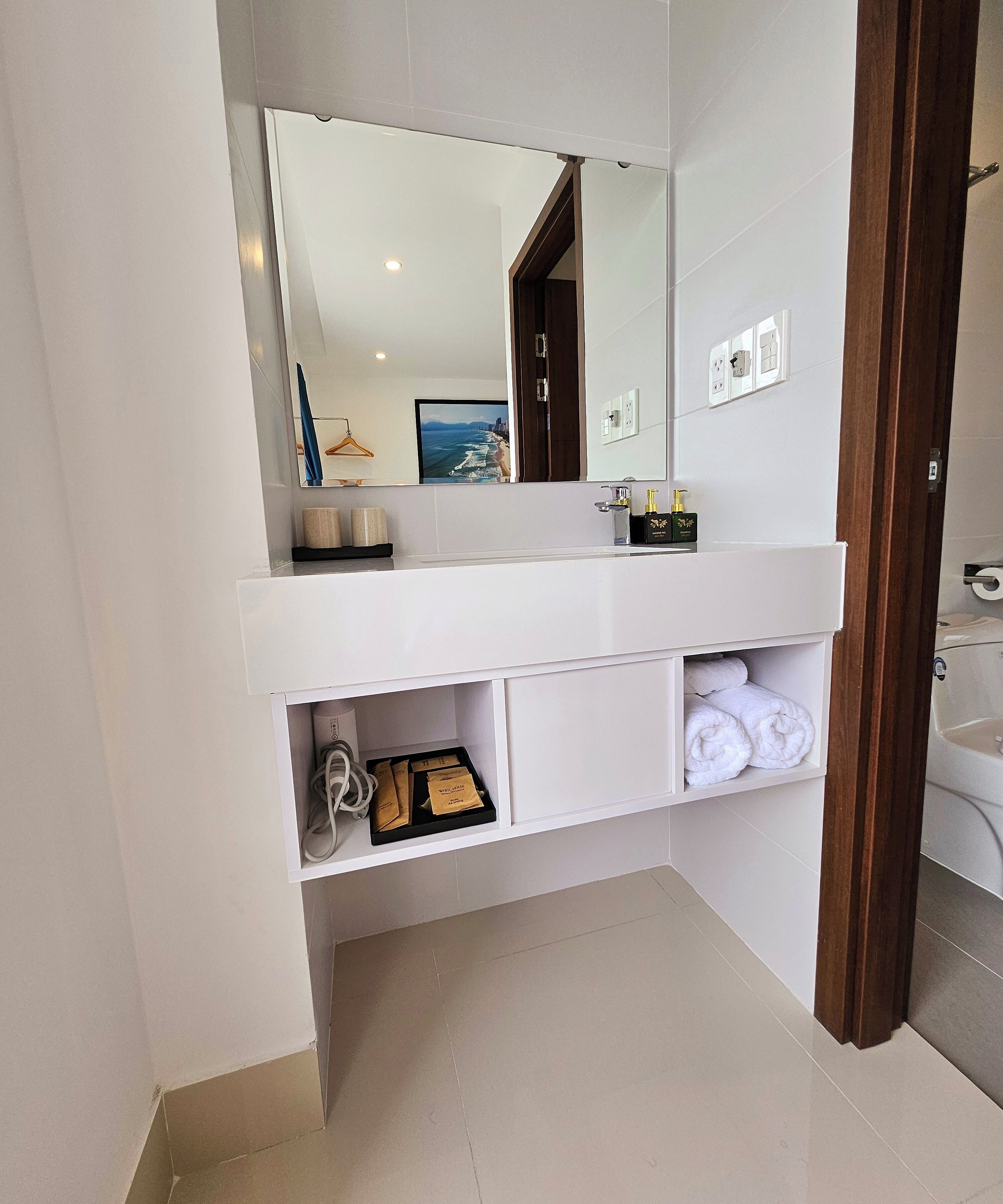 Un baño moderno con tocador blanco, un espejo grande y vista a la playa a través del espejo. Hay toallas y artículos de aseo en el tocador.
