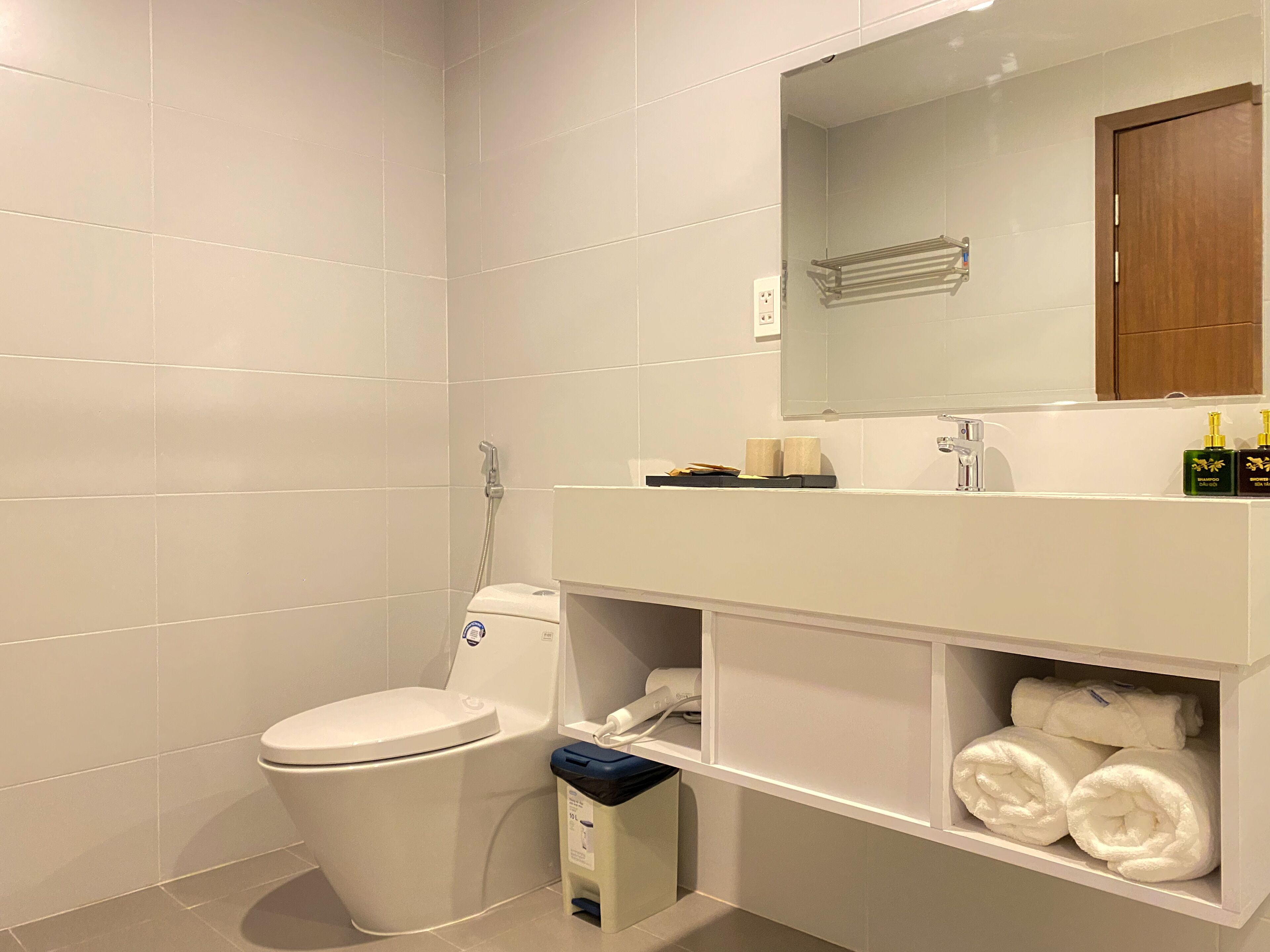 A modern bathroom with white tiles, a toilet, a sink with a mirror, and a shelf with towels.