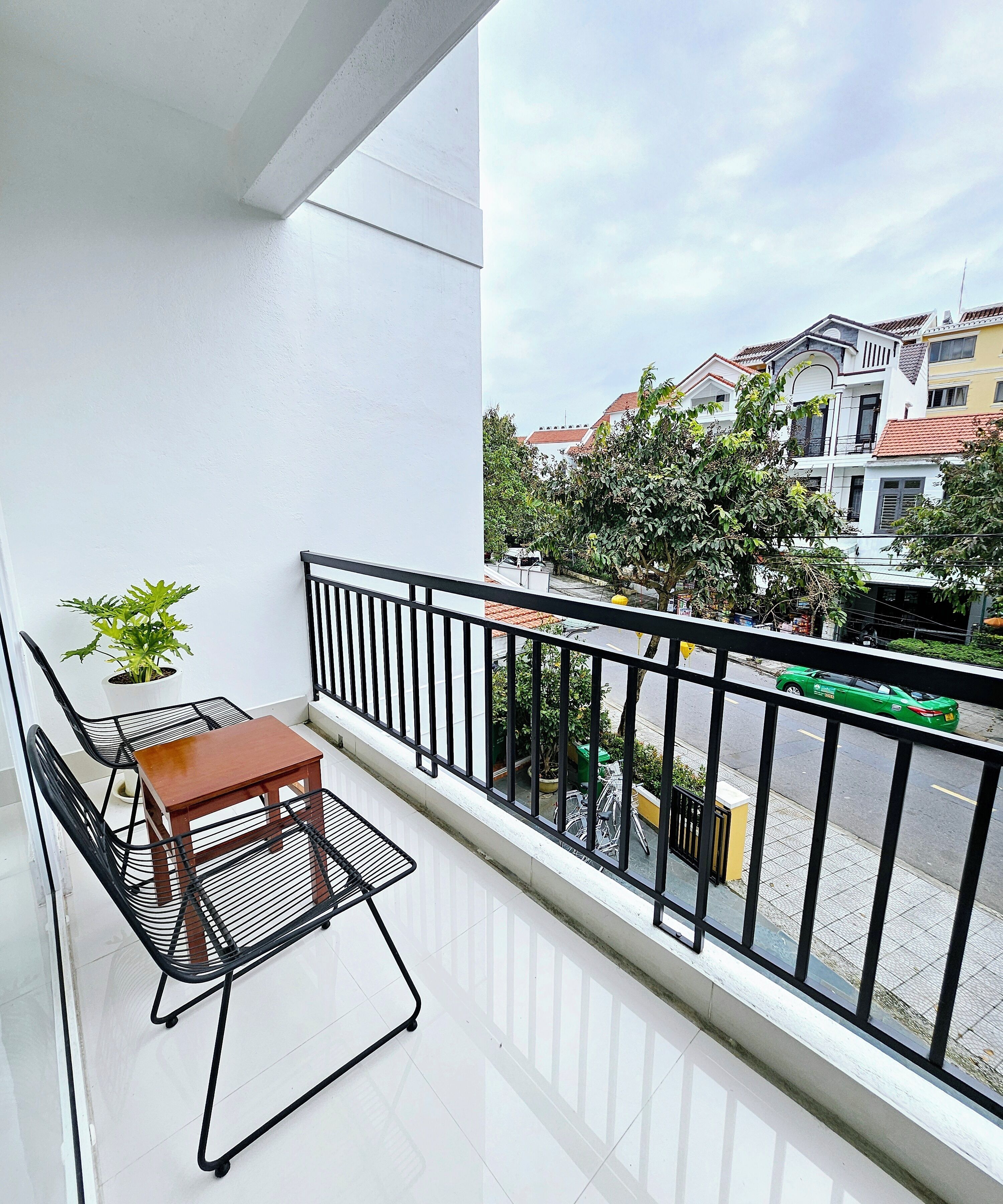A balcony with a table and chairs, a potted plant, and a view of a street with buildings and a green car.