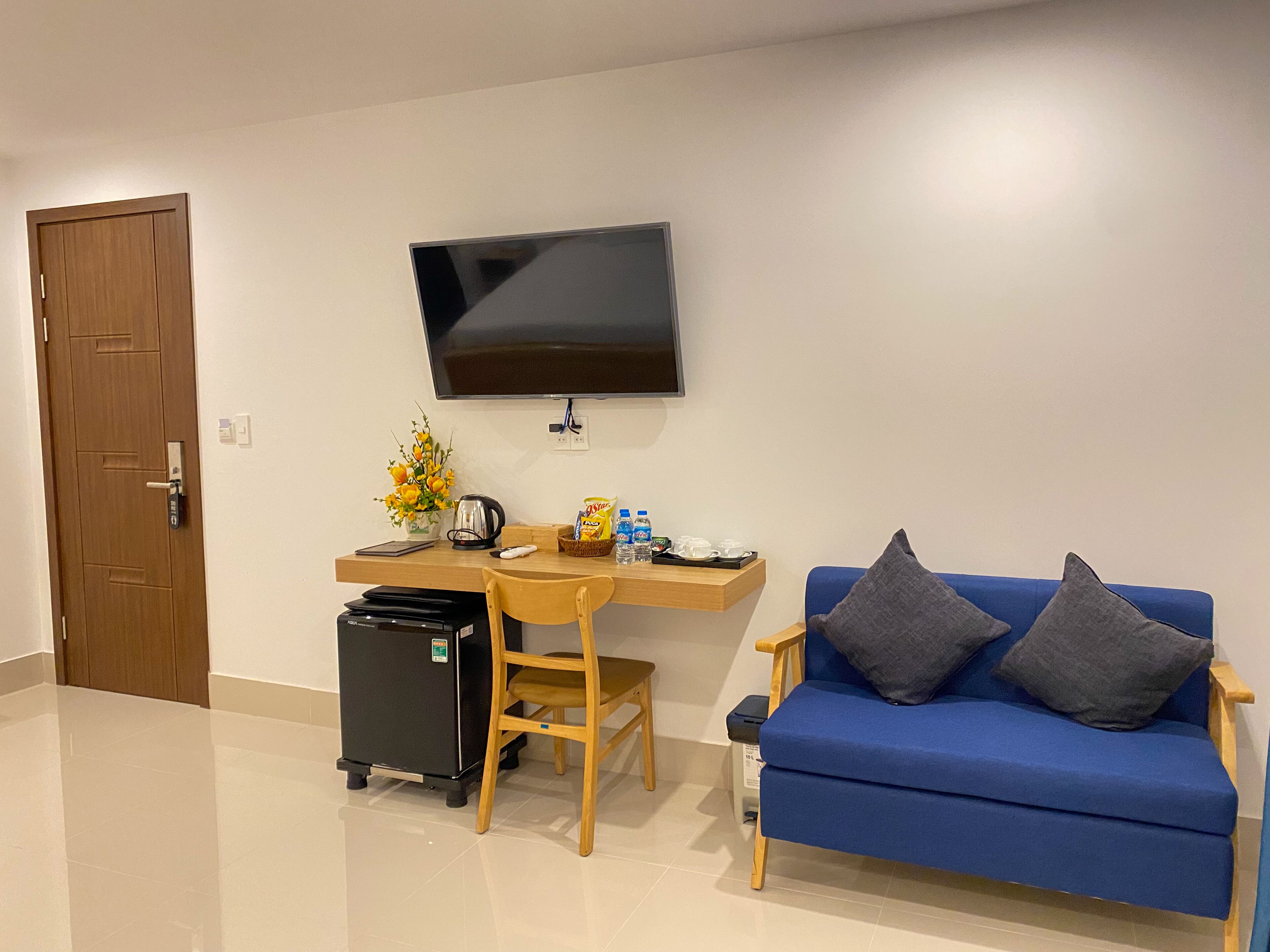 A room with a wooden desk, a blue sofa, a television mounted on the wall, a small table with a vase of flowers, and a black refrigerator.