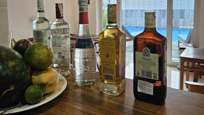 En bar med diverse flasker spiritus, en anretning med frugt og udsigt til en swimmingpool gennem store vinduer.