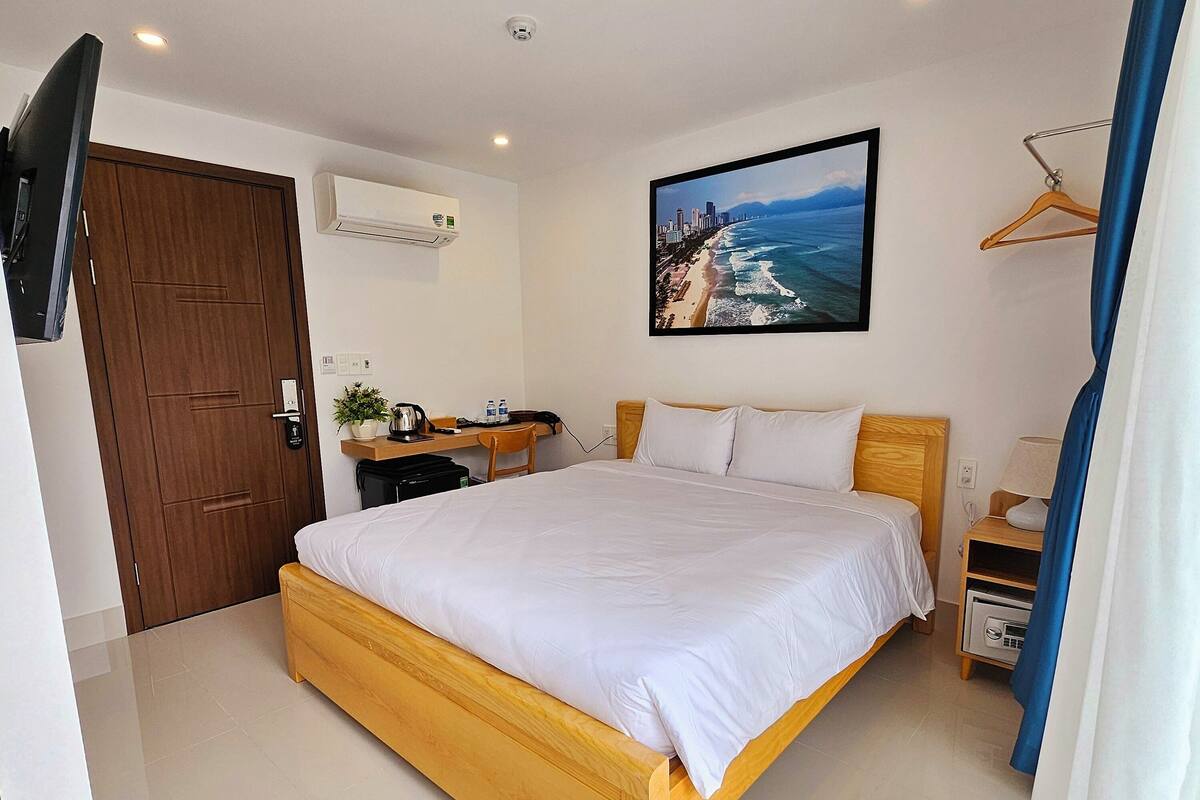 Ein Hotelzimmer mit einem großen Bett, einem Schreibtisch mit Stuhl, einem an der Wand befestigten Fernseher und einem Bild mit einer Strandansicht.