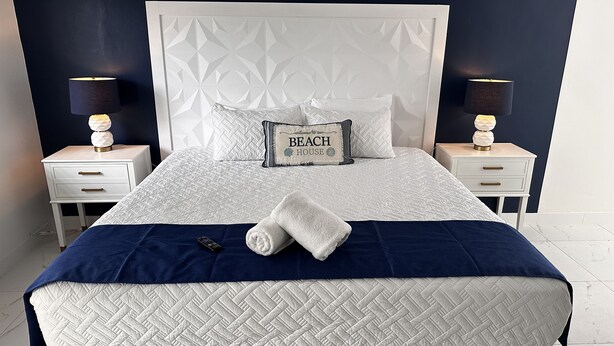 A neatly made bed with a white quilt, a blue blanket, and a pillow with "Beach House" text. There are two bedside tables with lamps and a small drawer.