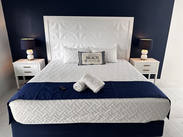 A neatly made bed with a white quilt, a blue blanket, and a pillow with "Beach House" text. There are two bedside tables with lamps and a small drawer.