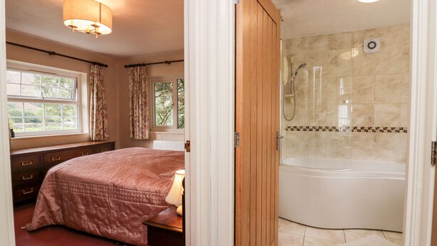 Een slaapkamer met een bed, nachtkastje, raam met gordijnen en een badkamer met bad en douche.