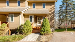 A two-story house with a front porch, a driveway, and a small front yard with two evergreen trees. - Cozy Dog Friendly Retreat w/ Hot Tub & Views (McHenry)