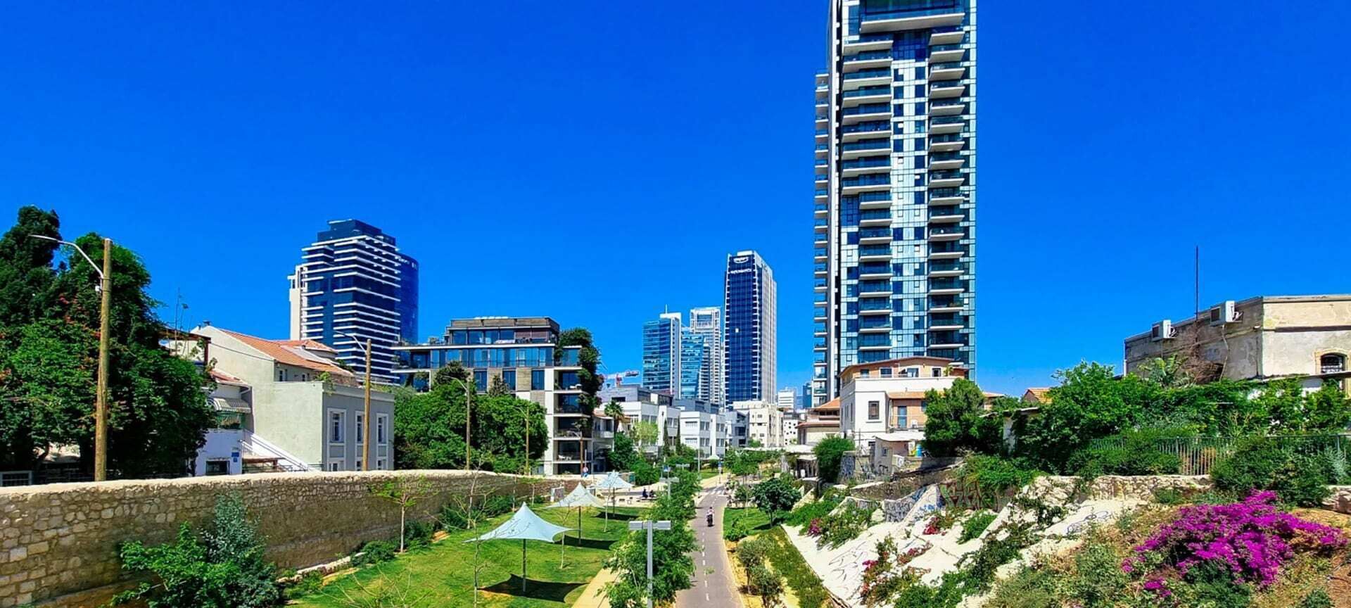 A cityscape with modern high-rise buildings, a clear sky, and a green park area with pathways.