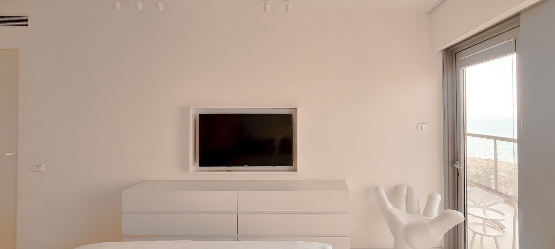 A bedroom with a bed, a TV mounted on the wall, a white chair, and a view of the sea through a large window.