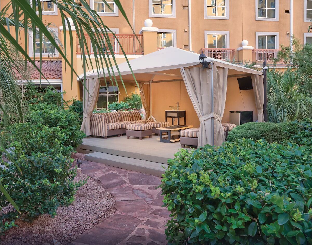 A covered outdoor seating area with sofas and a table, surrounded by greenery and a stone pathway.