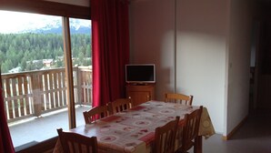 A dining area with a table set for four, a television on a stand, and a balcony with a view of a forest and mountains. - Equipped apartment in Superdévoluy (Le Devoluy)