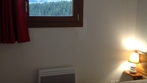 A hotel room with a bed, a window with a forest view, a heater, and a bedside lamp. - Equipped apartment in Superdévoluy (Le Devoluy)