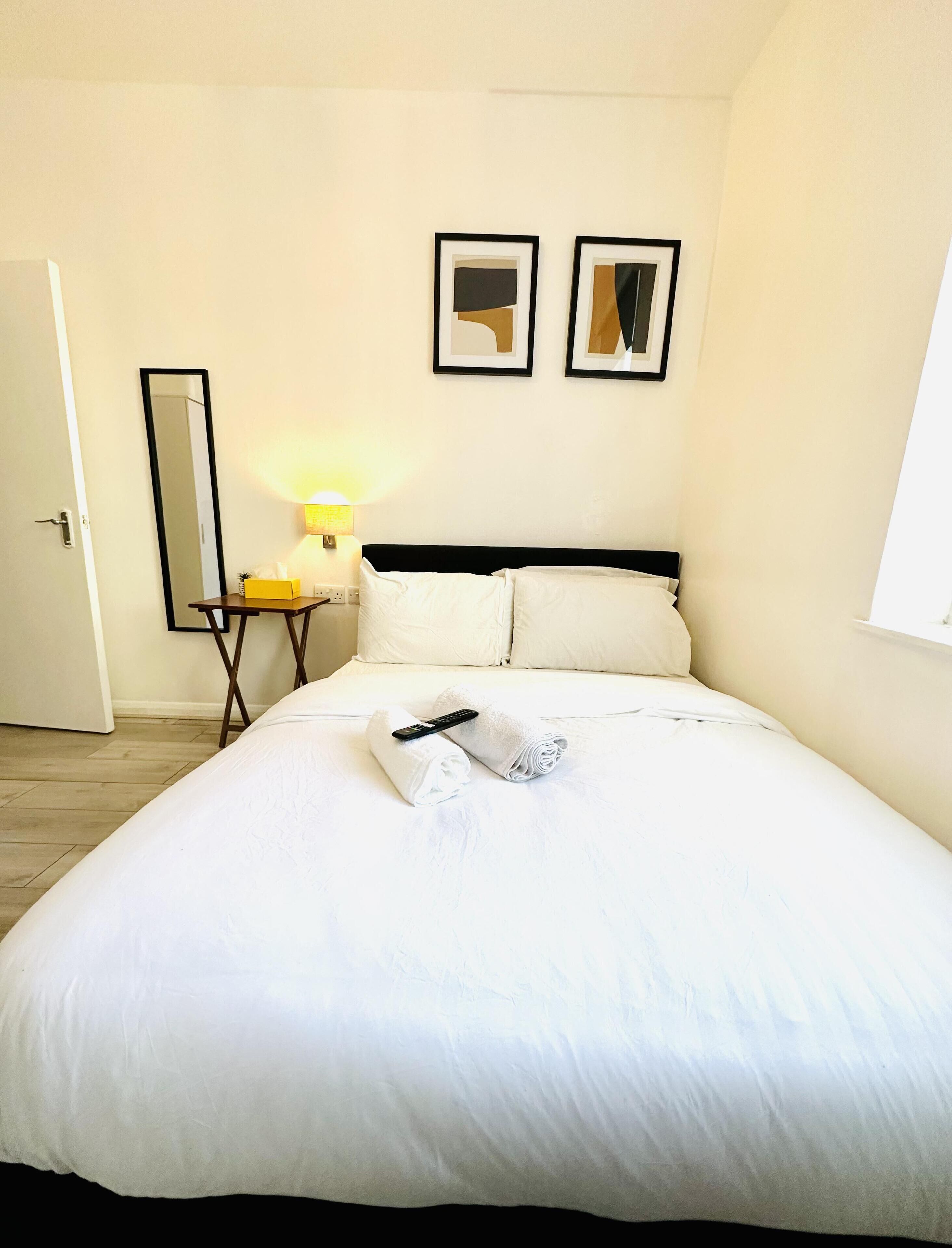 A neatly made bed with white linens, a remote control, and a rolled towel on a bedside table. There are two framed pictures on the wall above the bed.