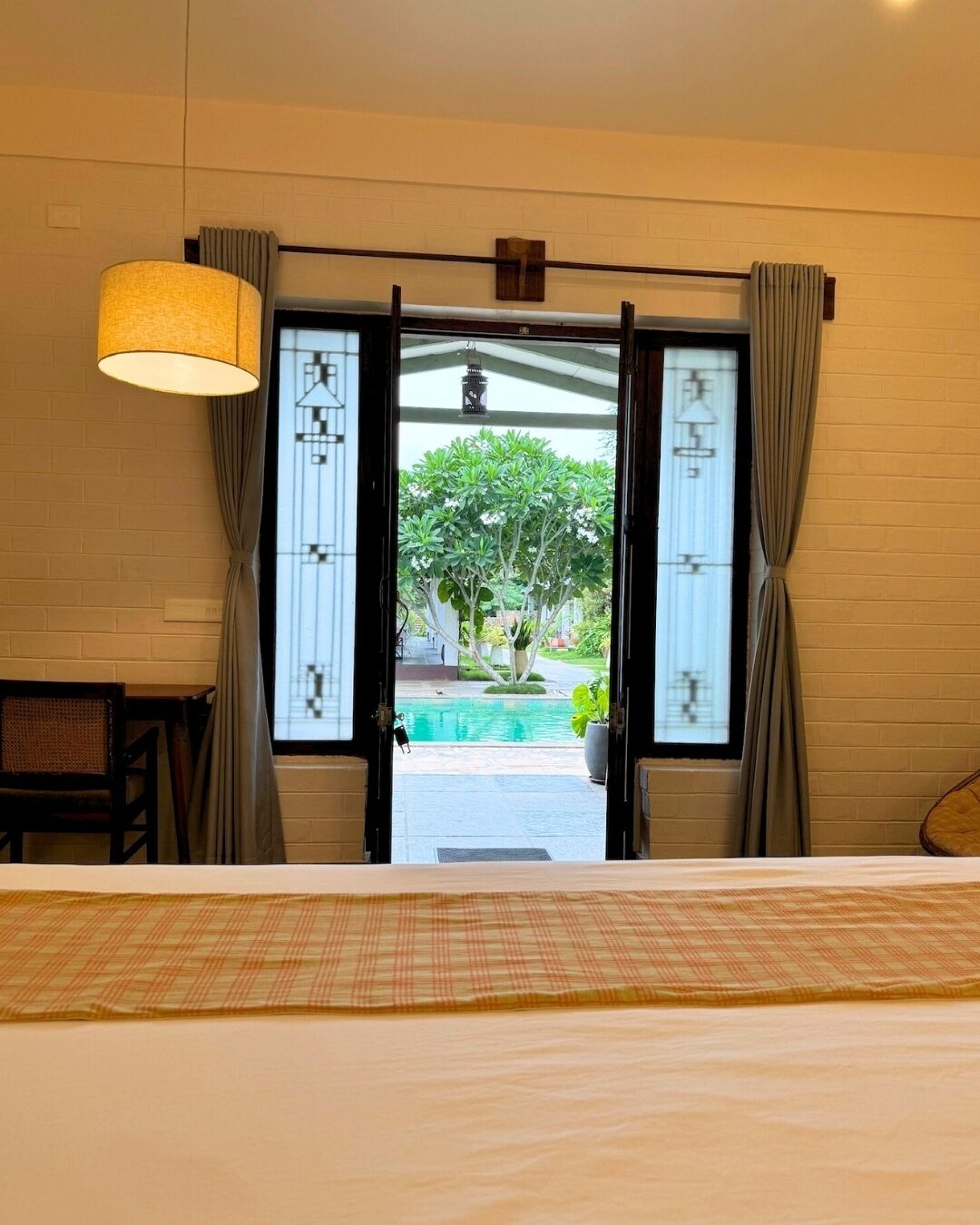 A bedroom with a bed, a desk, and a view of a swimming pool through open shutters.