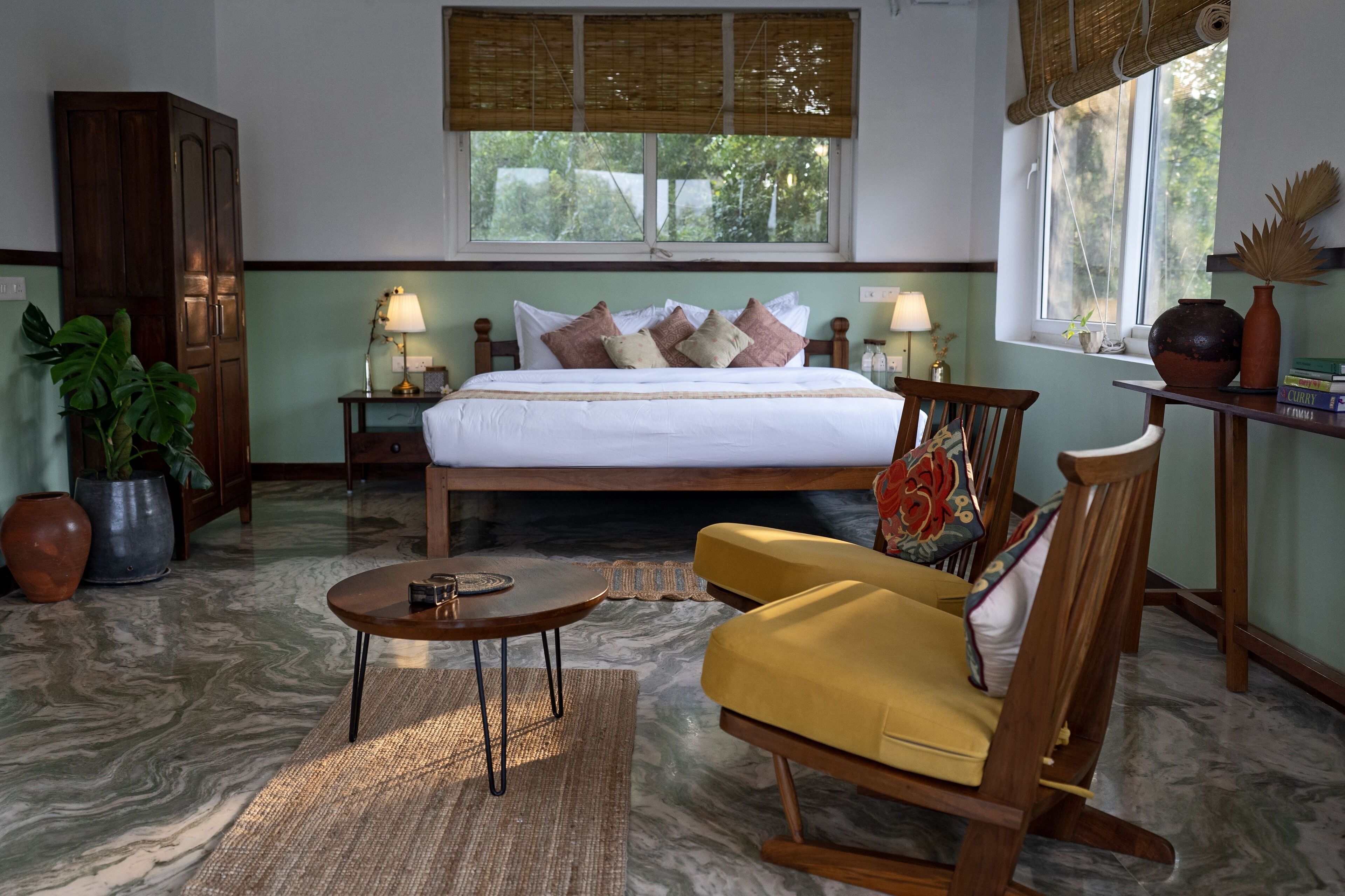 A bedroom with a bed, bedside tables, a wooden wardrobe, a round coffee table, a yellow sofa, and a window with a view of greenery.