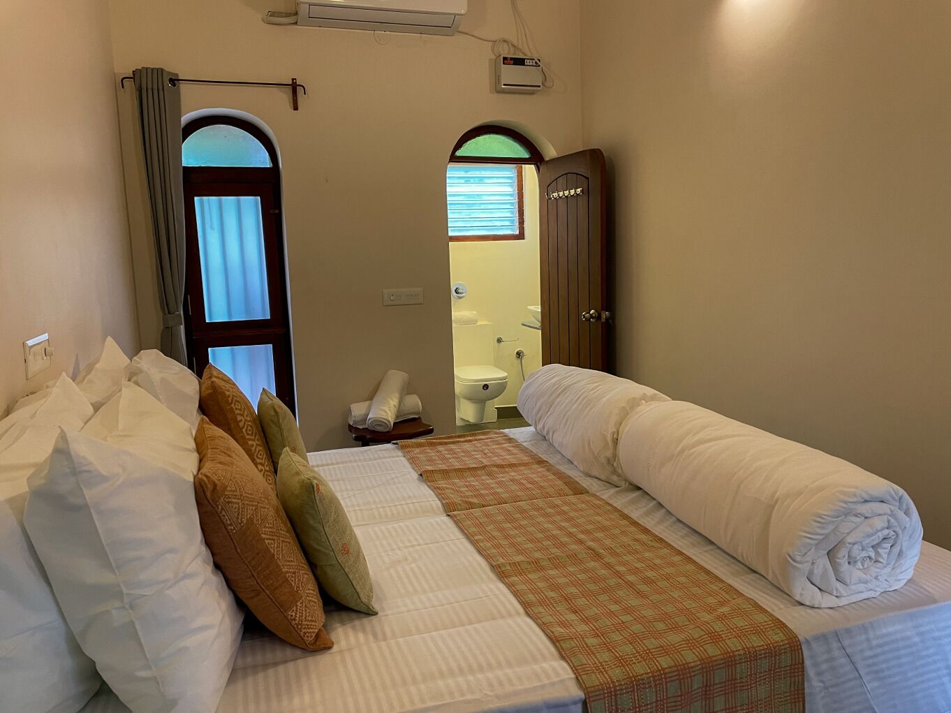 A hotel room with a bed, pillows, a blanket, a window with curtains, a door, and a bathroom visible through an open door.