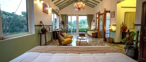 A spacious bedroom with a large bed, a sitting area with a sofa and armchairs, a dining table, and a view of the outdoors.