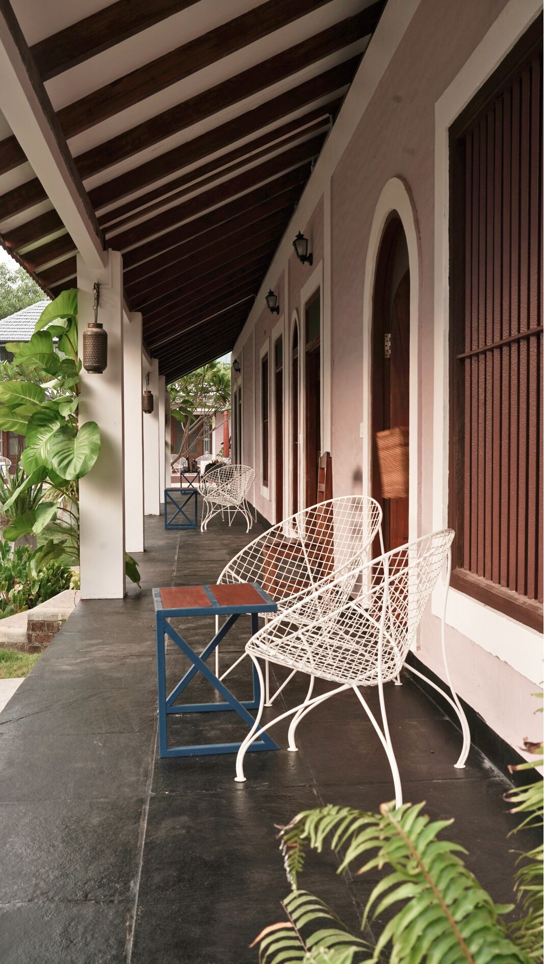 A covered outdoor seating area with white metal chairs and tables, surrounded by plants and a building with arched windows and a wooden ceiling.