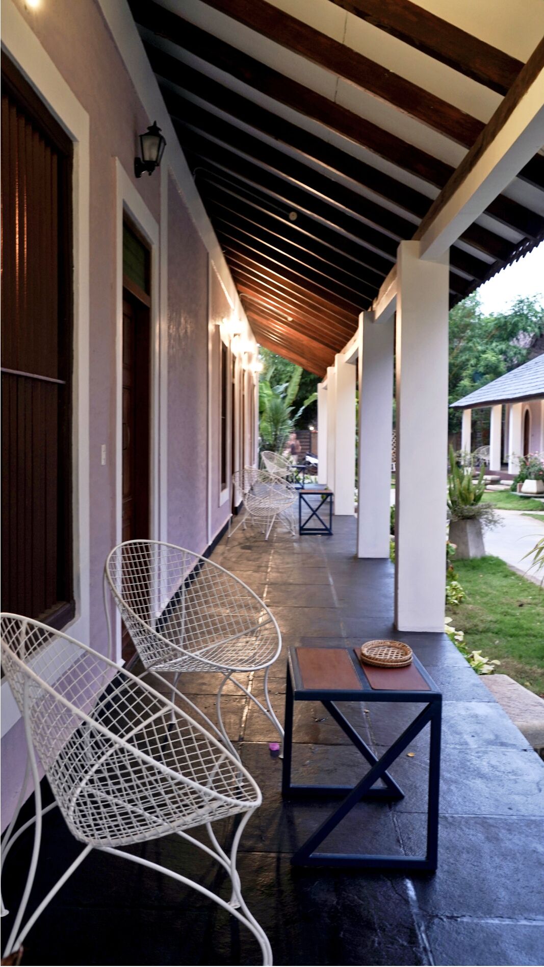 A covered outdoor area with white metal chairs and a small table, a wooden ceiling, and a view of a garden and house in the background.