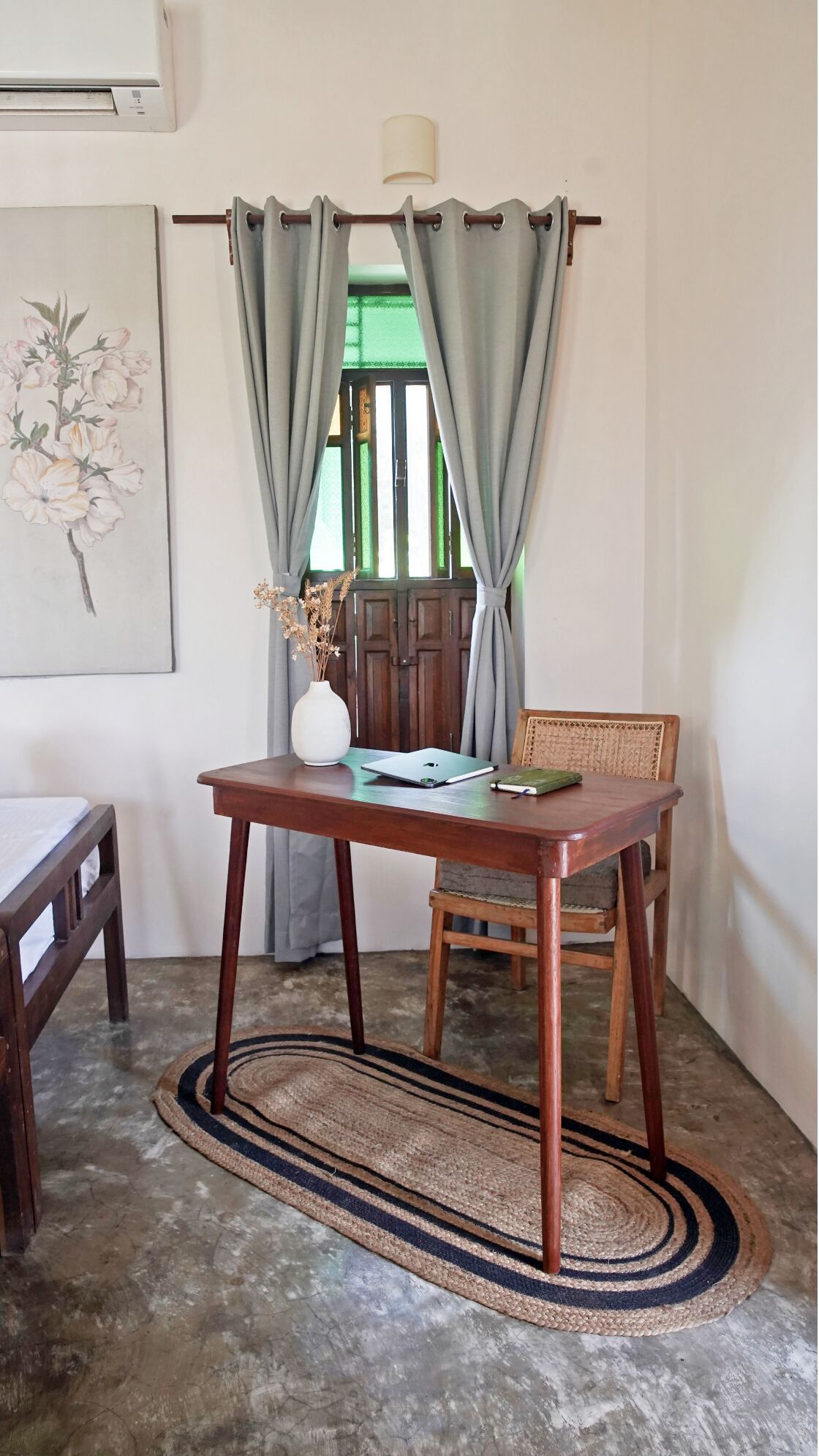 A room with a wooden table, a chair, a laptop, a notebook, and a vase with dried flowers.